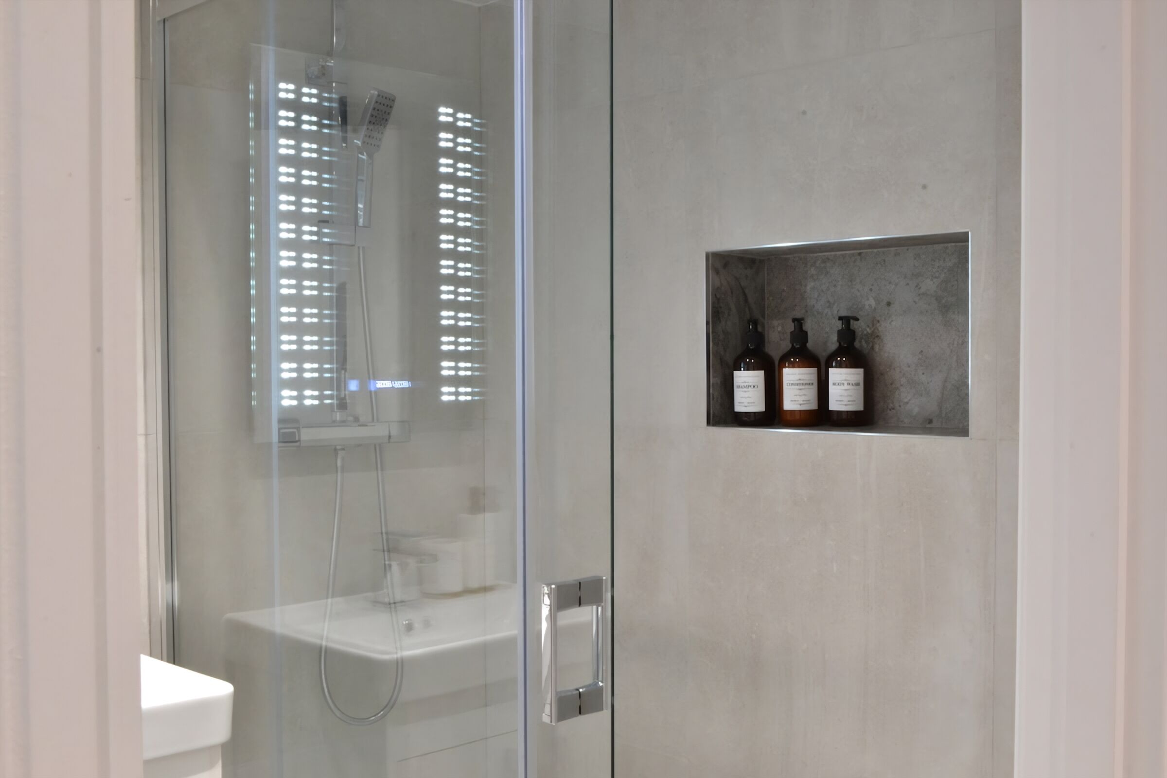 Une salle de bain moderne avec une douche à parois de verre, un lavabo blanc et une étagère sur laquelle sont posées trois bouteilles.