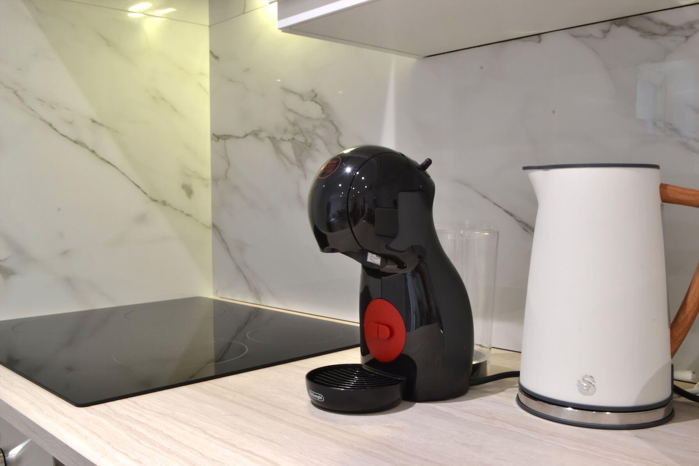 Une cuisine moderne avec une machine à café noire, une bouilloire électrique blanche et un récipient en verre posé sur un plan de travail en marbre.