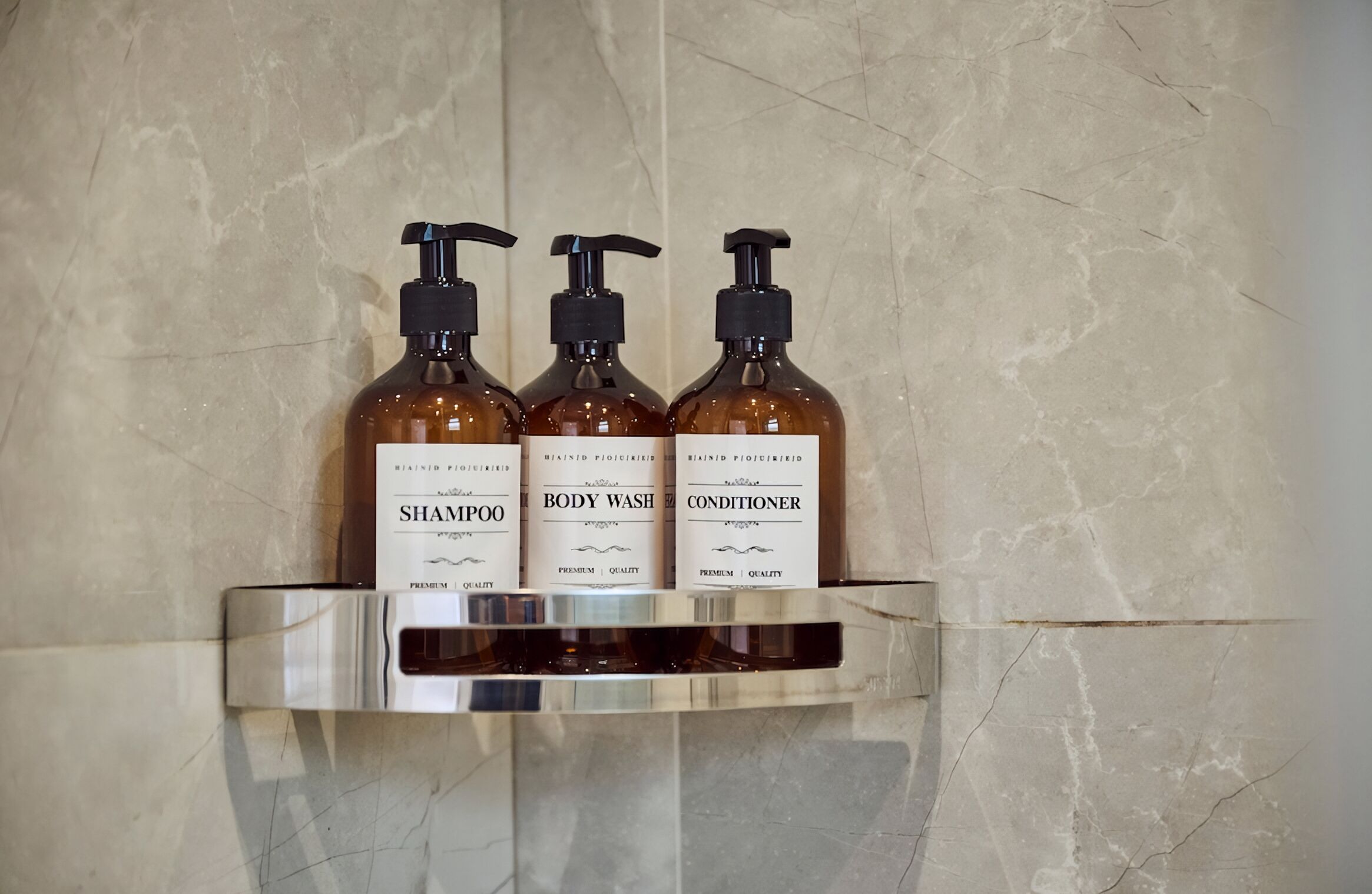 Drei Flaschen Duschgel, Shampoo und Conditioner auf einem Glasregal vor einer Marmorwand.