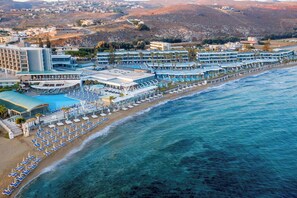 An aerial view of a coastal hotel with multiple buildings, a beach with rows of sun loungers, and a clear view of the ocean.