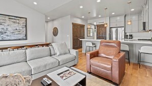 A modern living room with a grey sofa, a brown leather chair, and a coffee table. There is a kitchen area with grey cabinets and stainless steel appliances. - Moose Mgmt at Black Rock 14442: Fully Equipped Kitchen-Pvt Hot Tub-Dogs Welcome (Park City)