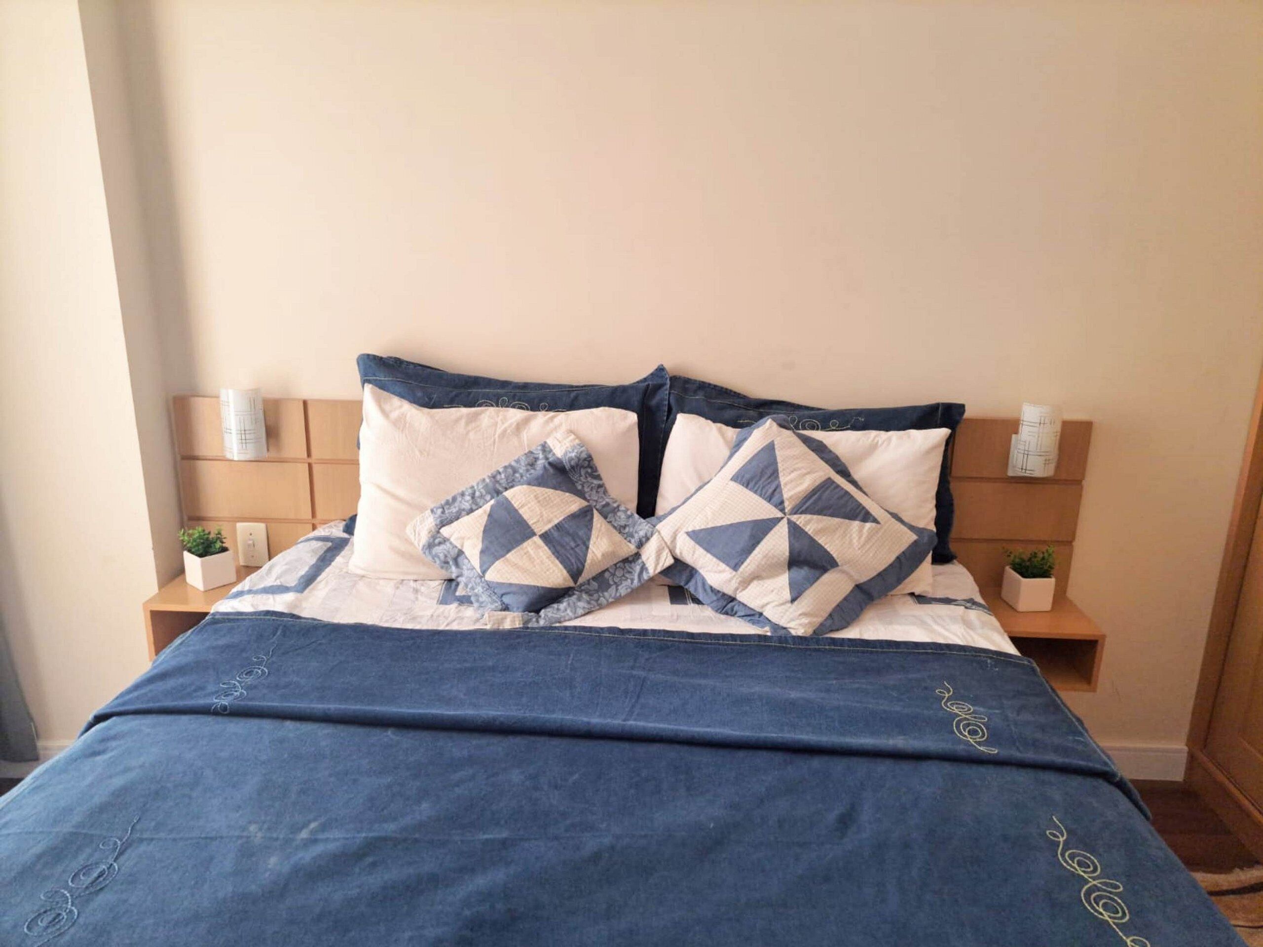 A neatly made bed with a blue and white bedspread, two decorative pillows, and a wooden nightstand with a small plant and a white container.