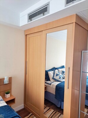 A bedroom with a wooden wardrobe, a bed with blue bedding, a small bedside table with a plant, and a radiator on the wall. - Get a Flat 1409 Mercury GRU (Guarulhos)
