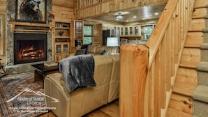 L’intérieur d’une cabane avec une cheminée en pierre, un canapé, un ventilateur de plafond et un coin cuisine visible à travers une porte ouverte.