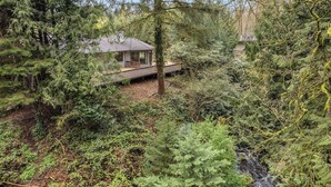 Une cabane dotée d’une terrasse en bois, entourée d’une forêt dense et d’une petite cascade.