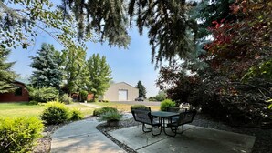 A patio with a table and chairs, surrounded by greenery and a building in the background.