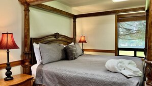 A four-poster bed with a grey quilt, two pillows, and a white towel on the bed. There are two bedside lamps with red shades.