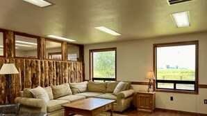 A living room with a sectional sofa, a wooden coffee table, a leather armchair, and a small cabinet. There are two windows providing a view of the outdoors.