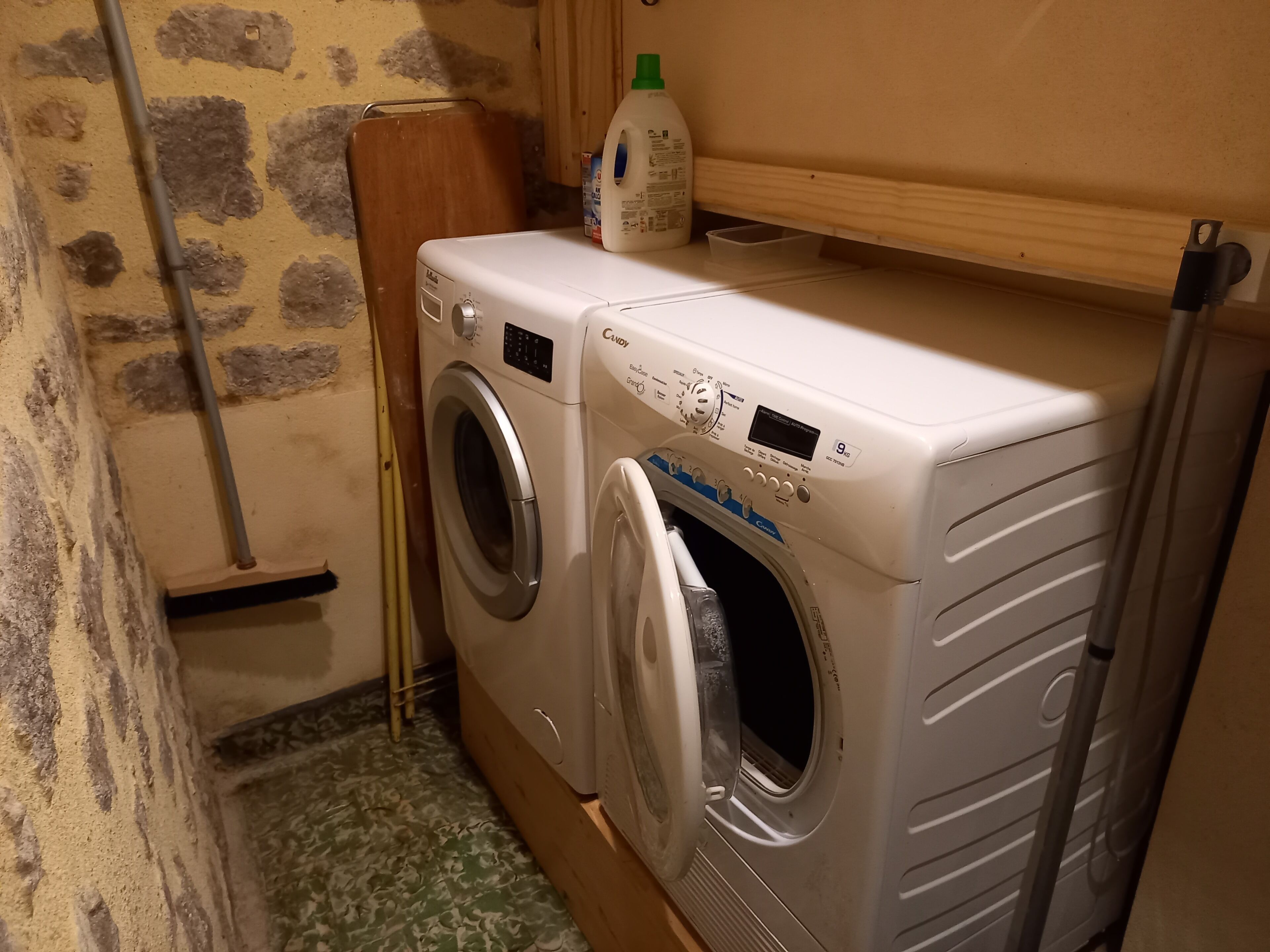 Una bugaderia petita amb dues rentadores frontals, un armari de fusta i una ampolla de detergent sobre una prestatgeria.