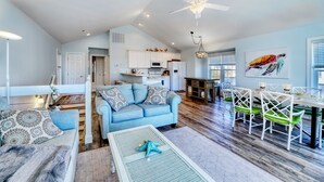 A spacious living area with a blue sofa, dining table, and chairs. There is a kitchen in the background with white cabinets and a ceiling fan. - Beach Bliss: 4br Beach Access Kayak Wi-fi 4 Bedroom Home by RedAwning (Holly Ridge)