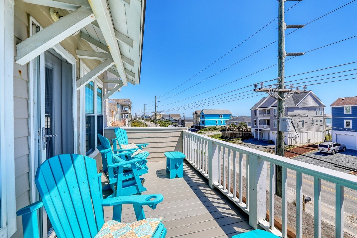 A wooden deck with blue Adirondack chairs and a small table, overlooking a residential area with houses and a clear blue sky.
