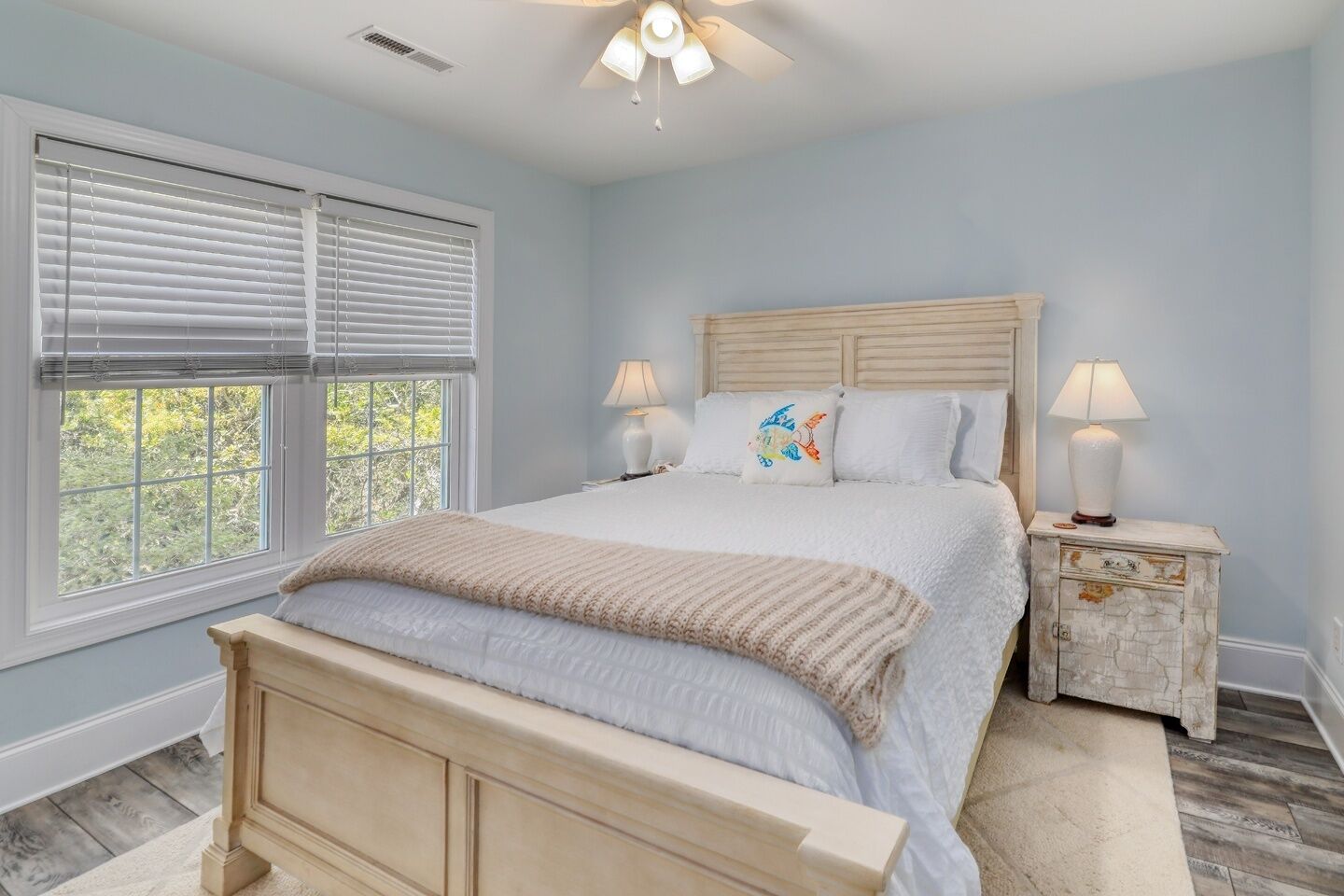 A bedroom with a bed, two bedside tables with lamps, a window with blinds, and a ceiling fan.
