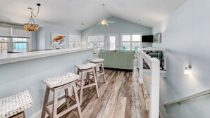 A spacious kitchen with a high ceiling, wooden flooring, and a central island. There are two bar stools with woven seats and a ceiling fan. - Beach Bliss: 4br Beach Access Kayak Wi-fi 4 Bedroom Home by RedAwning (Holly Ridge)