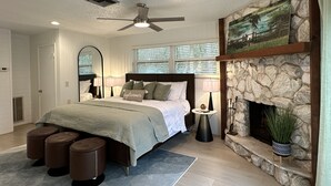 Un dormitorio con cama, ventilador de techo, chimenea de piedra y ventana con persianas.