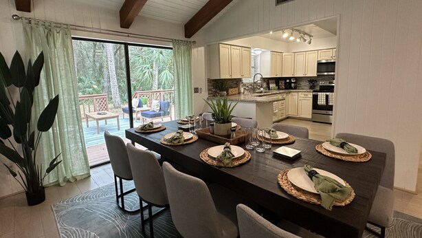 Un comedor moderno con una mesa grande preparada para cuatro personas, rodeada de sillas. Se aprecia una terraza y vegetación al exterior.