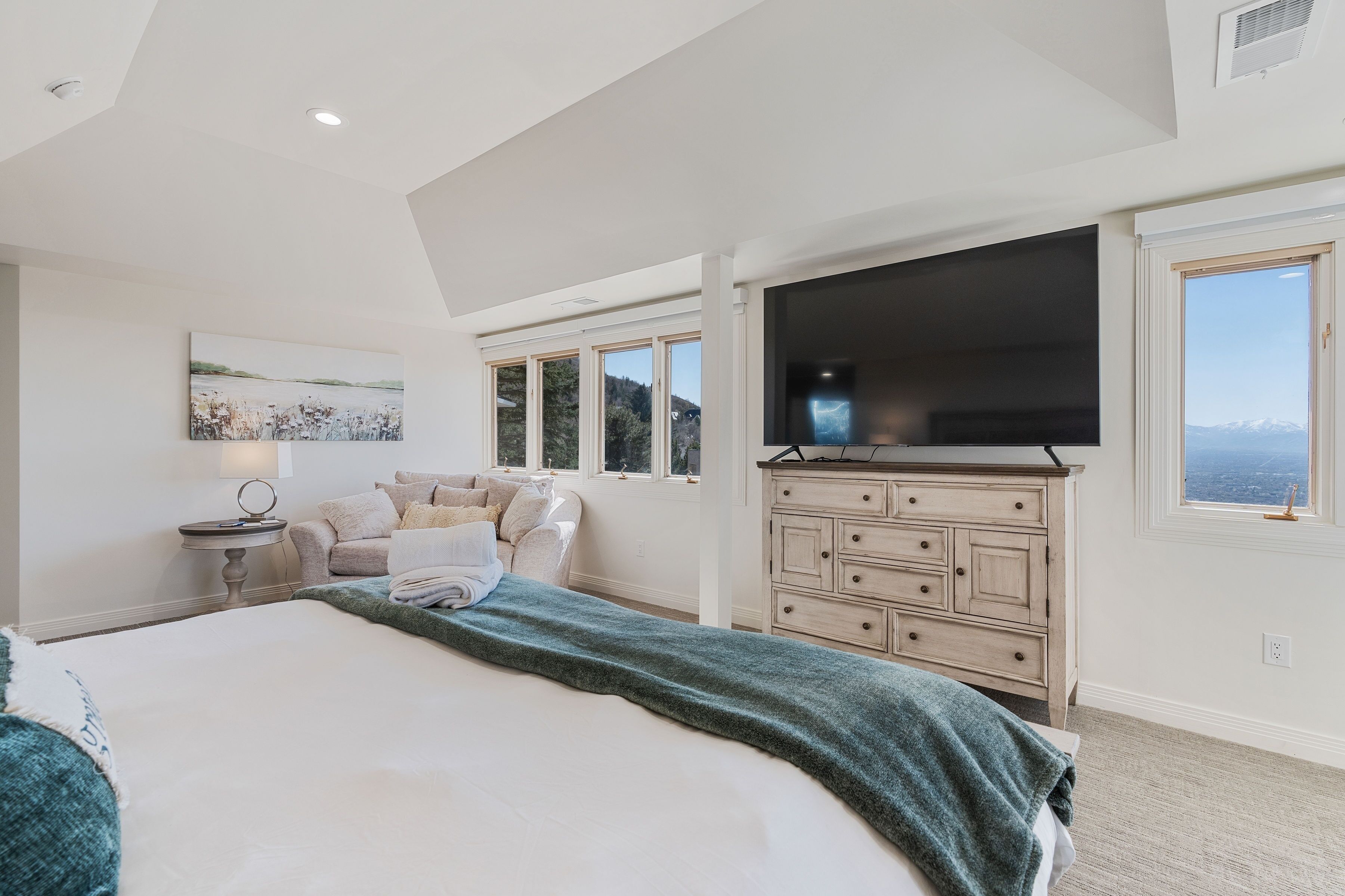 A bedroom with a large bed, a flat-screen TV mounted on the wall, a dresser, and a window with a mountain view.