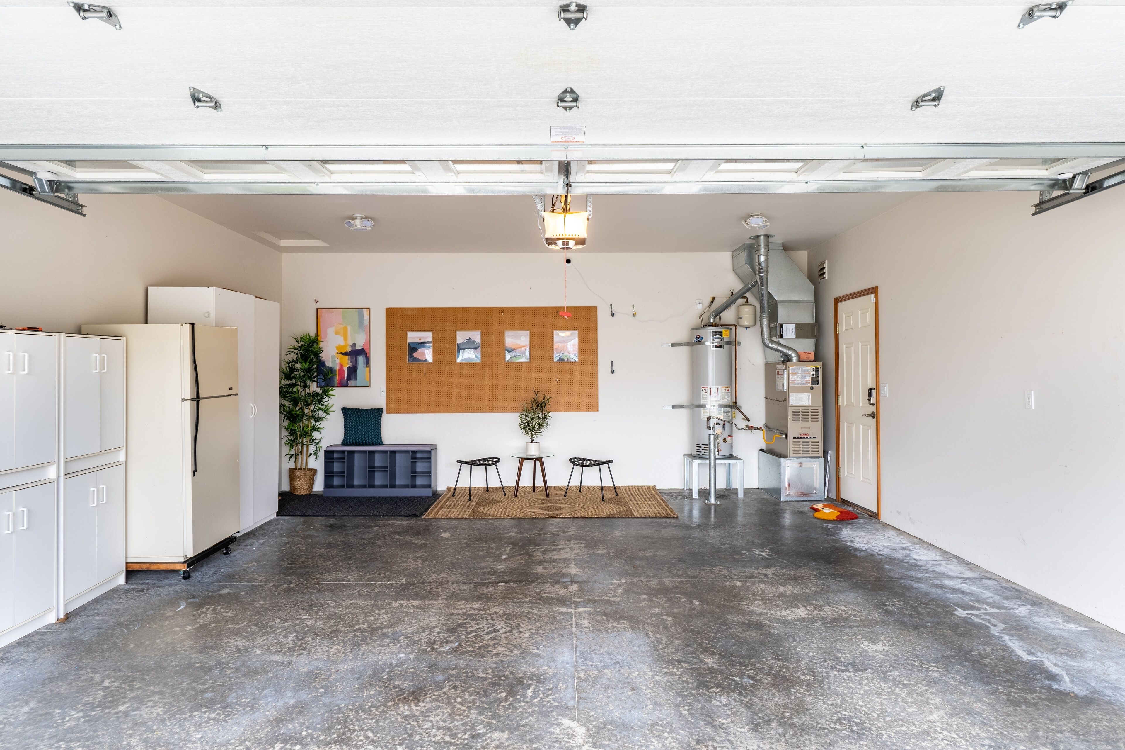 Een ruime garage met een betonnen vloer, een koelkast, kastjes, een tafel met stoelen, een potplant en een wandlamp.