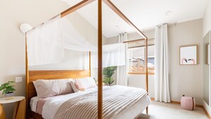 A four-poster canopy bed with a wooden headboard, a bedside table with a plant, and a large window with curtains.