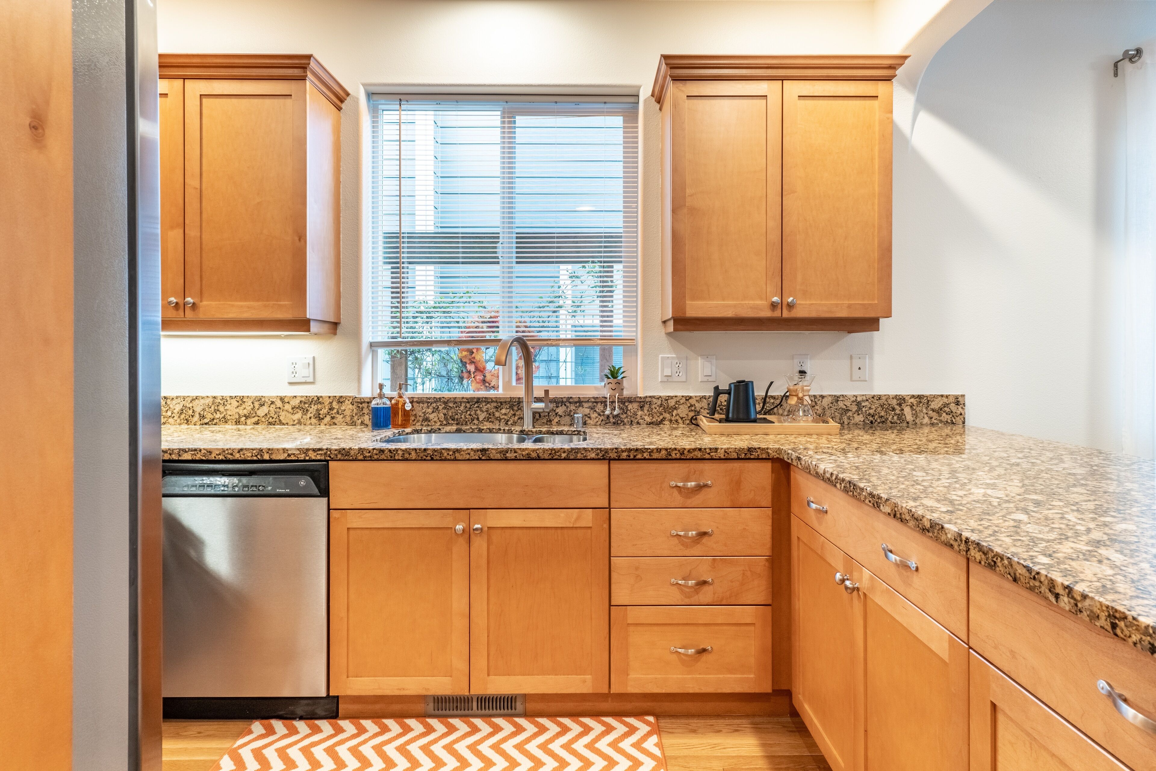 A kitchen with wooden cabinets, a granite countertop, a dishwasher, and a window with blinds.