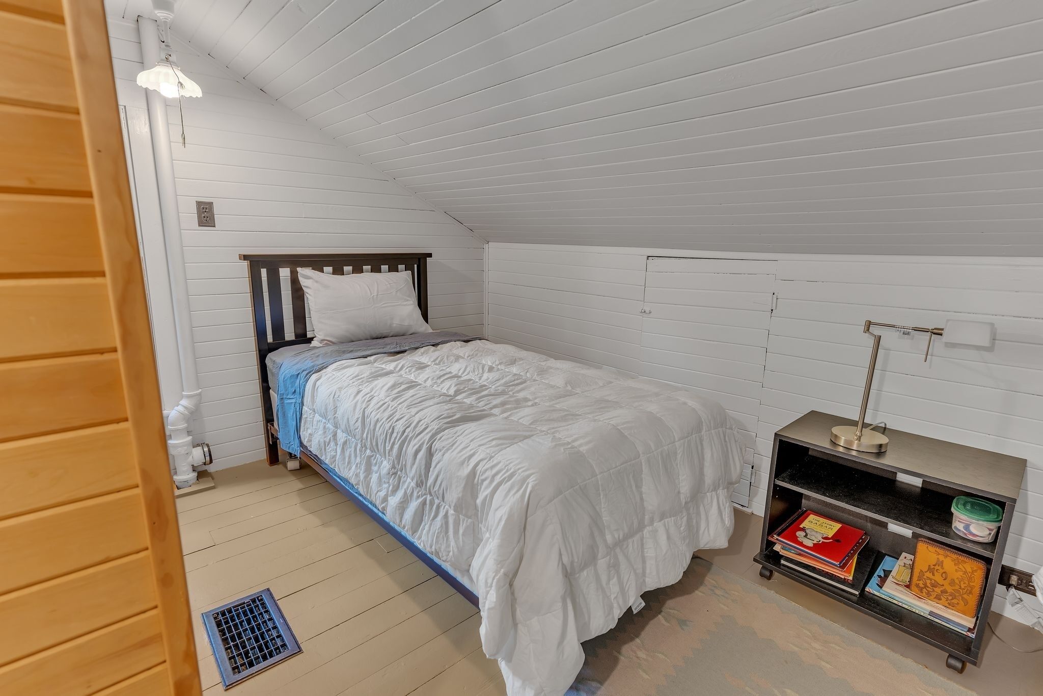 A bedroom with a bed, a nightstand, a shelf with books, and a window.