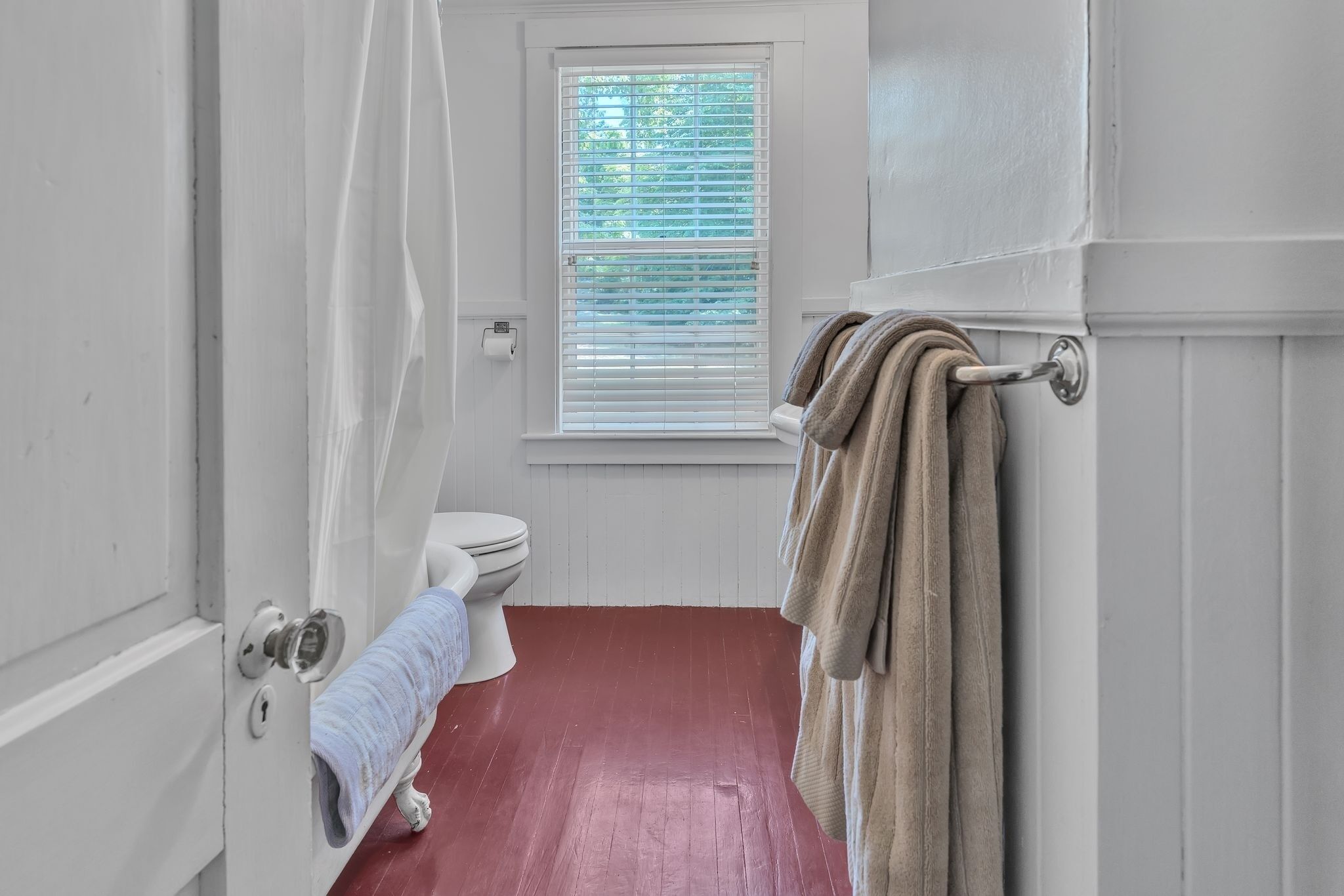 A bathroom with a white toilet, a window with blinds, a red floor, and a towel rack with towels.
