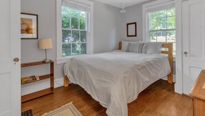 Une chambre avec un grand lit, deux fenêtres, une table de chevet avec une lampe et un plancher en bois.