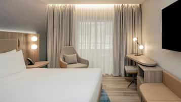 A modern hotel room with a large bed, a desk with a chair, a seating area, and a TV mounted on the wall.