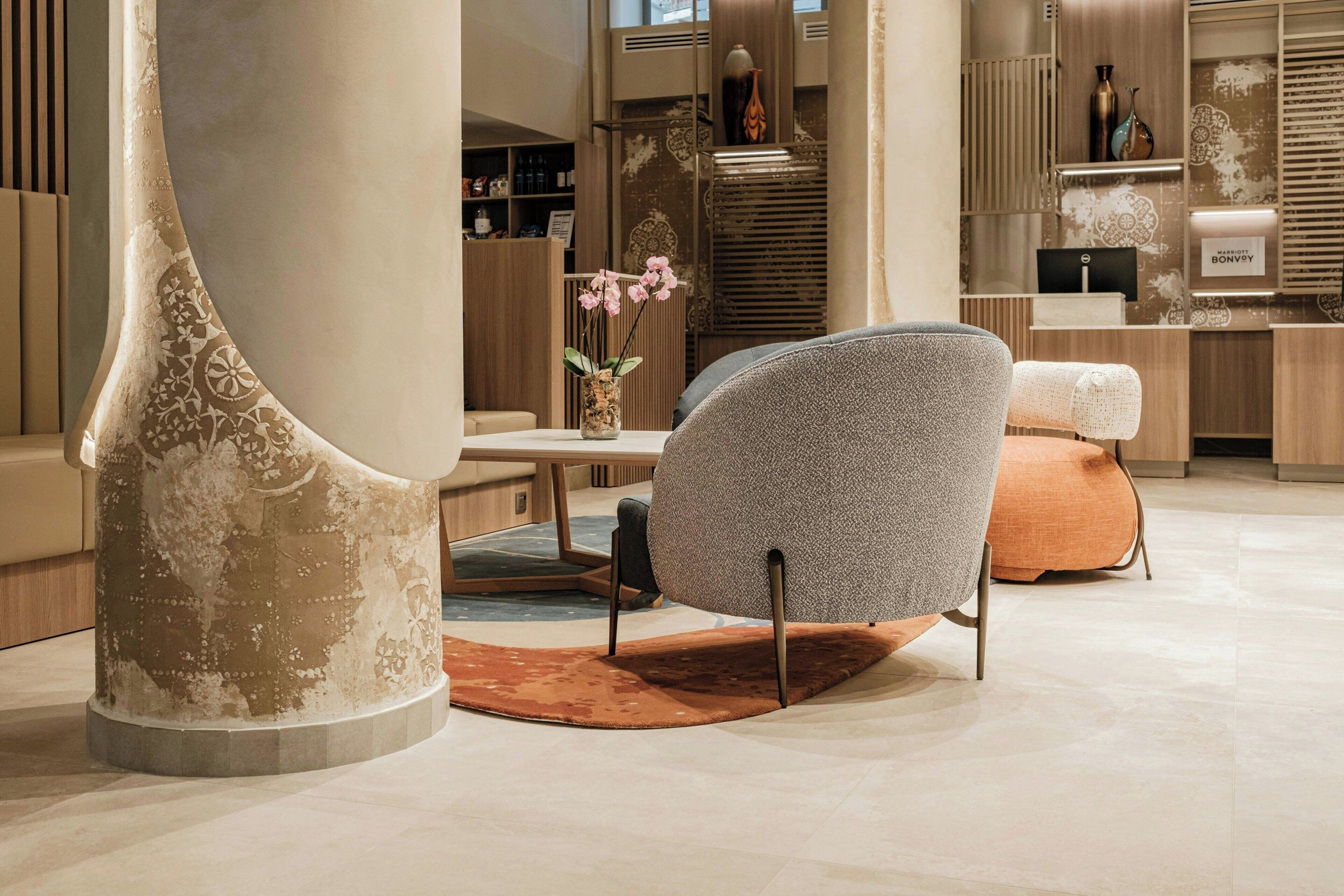 A modern interior with a unique curved column, a grey upholstered chair, a small orange ottoman, and a vase with pink flowers.