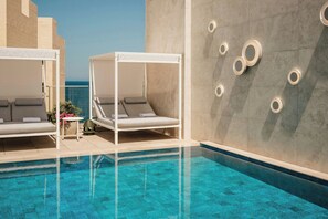 Área à beira da piscina com duas camas de dossel brancas, uma mesa pequena com um vaso de flores e uma parede com luminárias circulares.