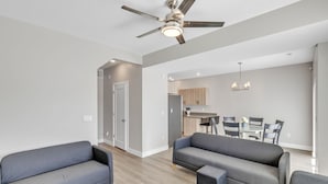 A modern living room with grey sofas and a ceiling fan, leading to a kitchen area with wooden cabinets and a dining table. - The Ideal Getaway 3 Bedroom Home by RedAwning (Cedar City)