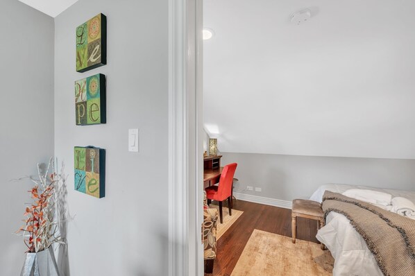 A modern bedroom with a bed, a desk, and a chair. There are wall art pieces and a vase with flowers.