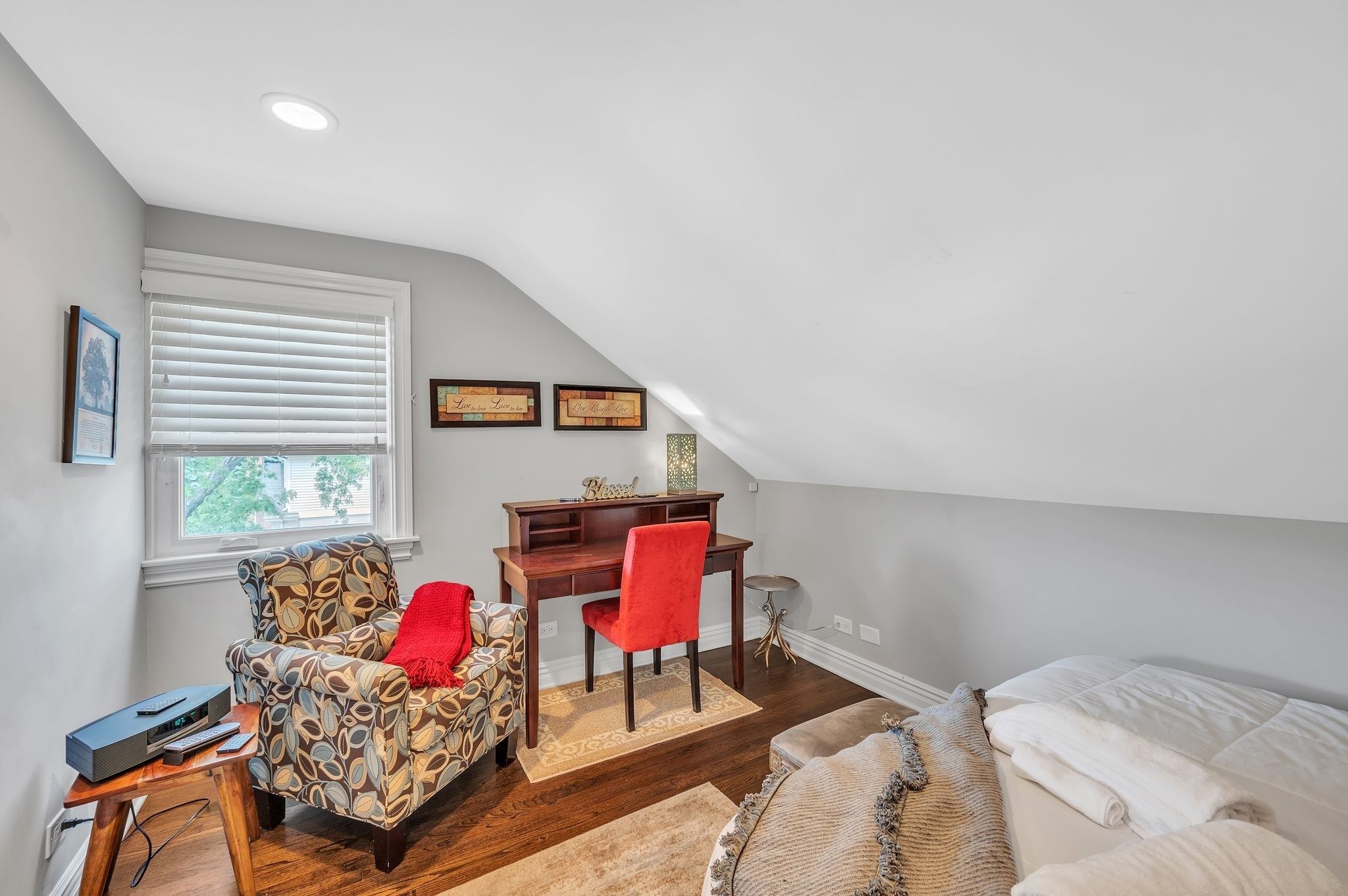 A cozy attic bedroom with a sloped ceiling, a desk with a red chair, a patterned armchair, and a bed with a white comforter.
