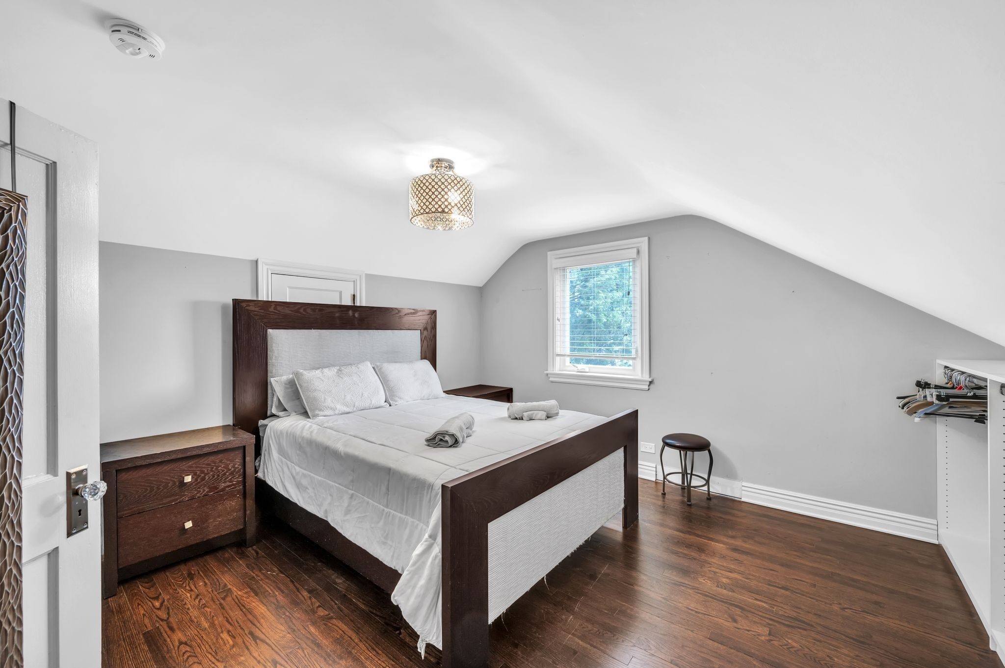Une chambre à coucher avec un lit en bois, une table de chevet, un petit tabouret, une fenêtre avec des stores et un luminaire au plafond.