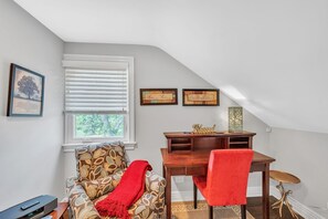Una habitación acogedora con ventana, una silla con una manta roja, un escritorio de madera y una mesita pequeña.