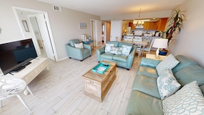 A living room with a TV, sofas, a coffee table, and a kitchen in the background. - Beach House 503d 2 Bedroom Condo by RedAwning (Miramar Beach)