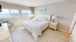 A bedroom with a large bed, a nightstand, a chair, and a view of the sea through the windows. - Beach House 503d 2 Bedroom Condo by RedAwning (Miramar Beach)