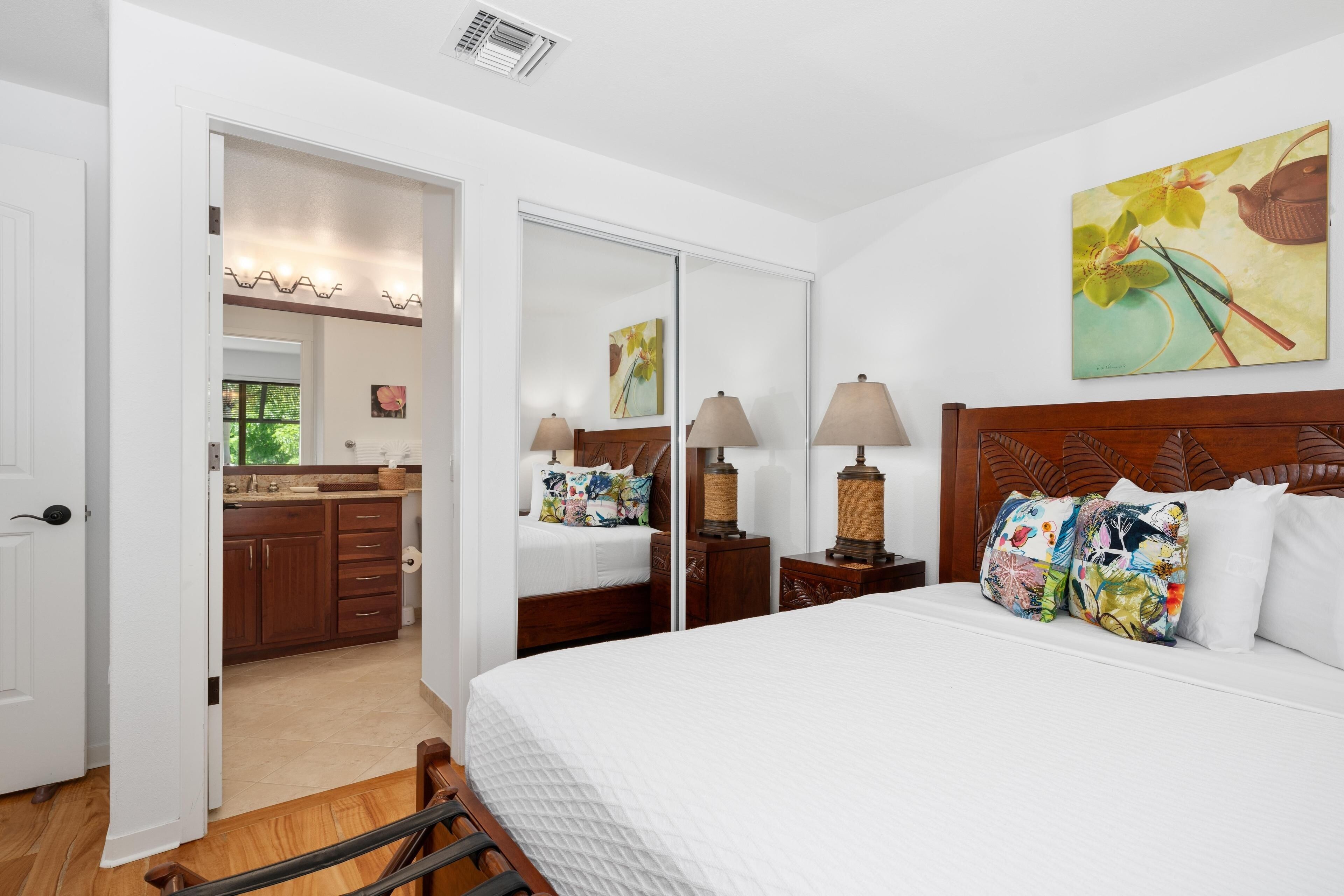 A hotel room with a bed, bedside lamps, a wooden headboard, a painting, and a bathroom visible through an open door.