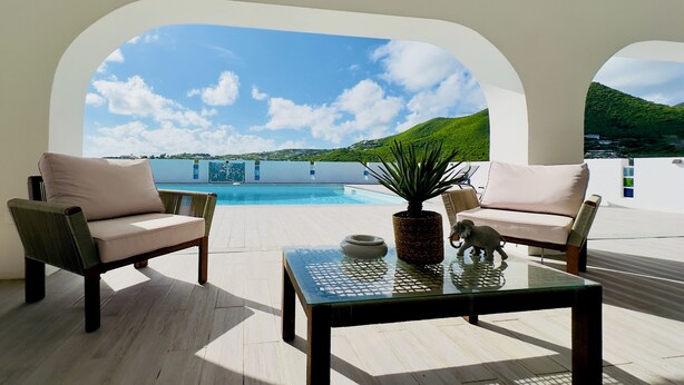 A sunlit patio with two cushioned chairs, a glass coffee table, and a view of a pool and green hills. - Charming spacious 3-bedroom villa with panoramic views of Orient Bay Beach.

 (Mont Vernon)