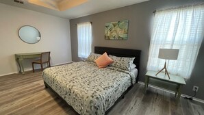 Un dormitorio con cama, escritorio, silla, lámpara y un cuadro en la pared.