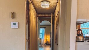 Un pasillo con suelo de madera, un candelabro y vistas a una habitación con ventana y cortinas.