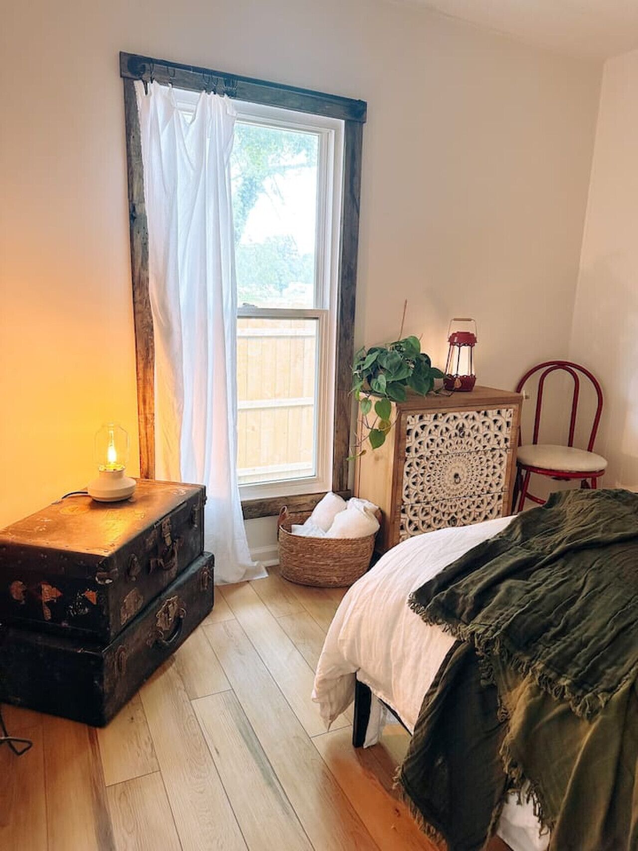 Een slaapkamer met een houten vloer, een bed met een groene deken, een vintage koffer, een rieten mand, een decor kast en een raam met witte gordijnen.