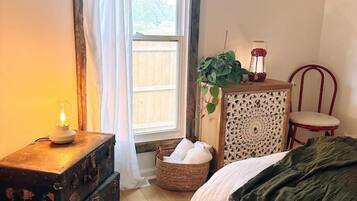 Een slaapkamer met een houten vloer, een bed met een groene deken, een vintage koffer, een rieten mand, een decor kast en een raam met witte gordijnen.