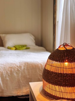 Una cama con colcha y almohadas blancas, una lámpara de mimbre sobre una mesita de noche y una ventana con cortinas.