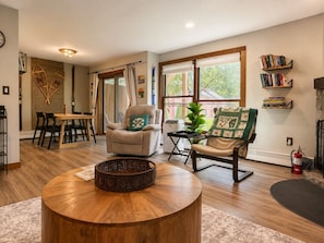 A spacious living room with a round wooden coffee table, two armchairs, a dining area with a wooden table and chairs, and a bookshelf.
