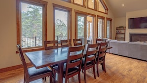 A dining area with a wooden table and chairs, large windows with a view of trees, and a fireplace. - Beautiful New Home with Views of Castle Mountain - 3510 (Estes Park)