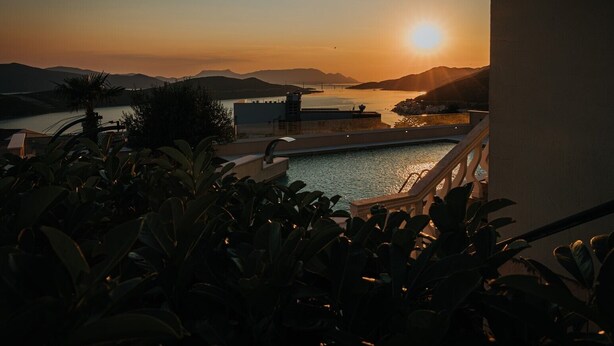 A pool with a sunset view, surrounded by greenery and overlooking a body of water and distant hills.