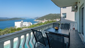 A balcony with a table and chairs, overlooking a clear blue sea and a town in the distance.