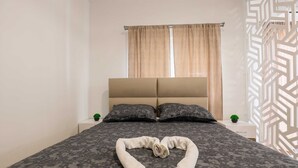 A neatly made bed with a heart-shaped towel on the bed, two pillows, and a nightstand with a small plant.