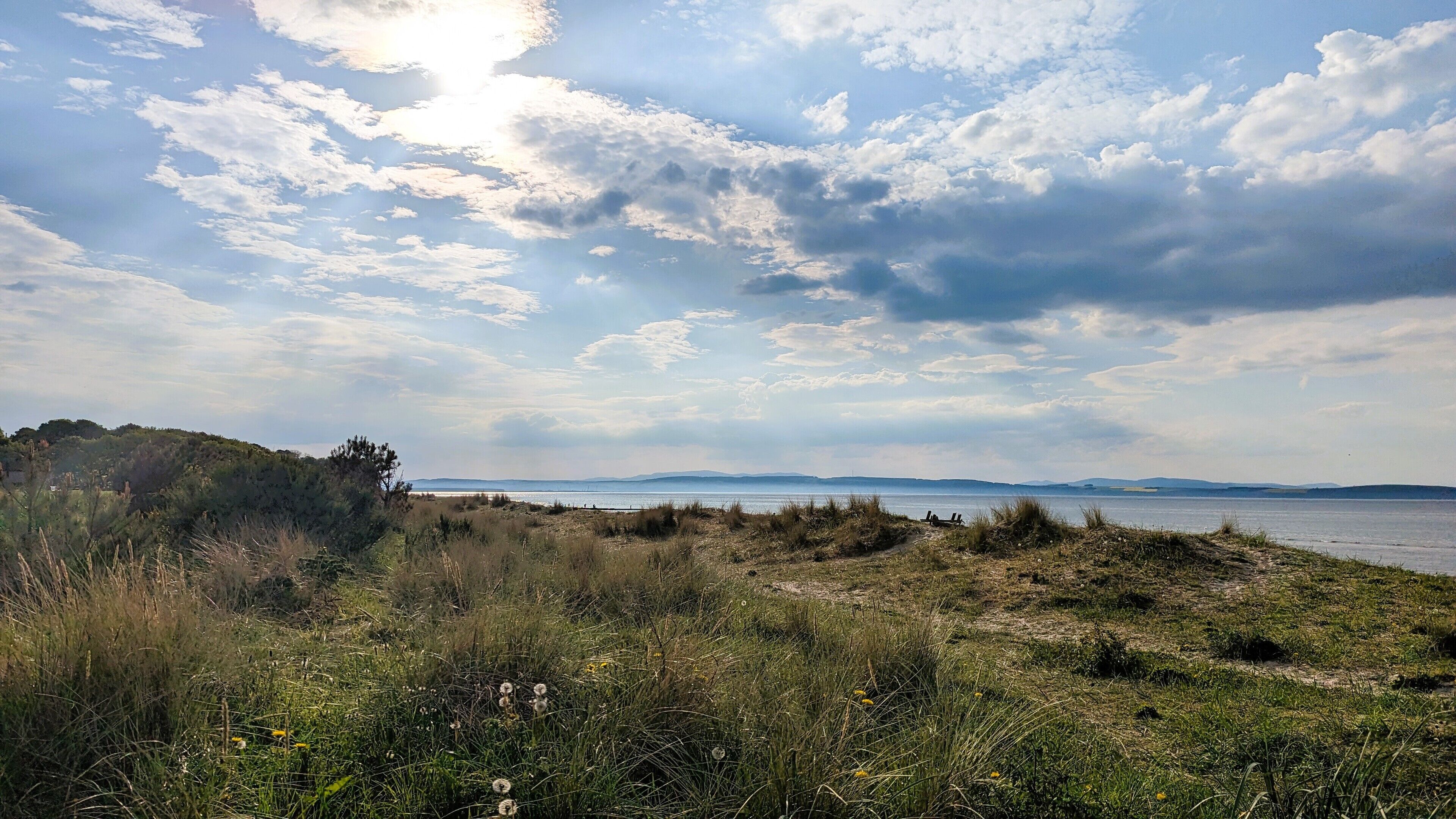Eine Küstenlandschaft mit einer grasbewachsenen Vordergrundfläche, einem Gewässer und fernen Hügeln unter einem teilweise bewölkten Himmel.