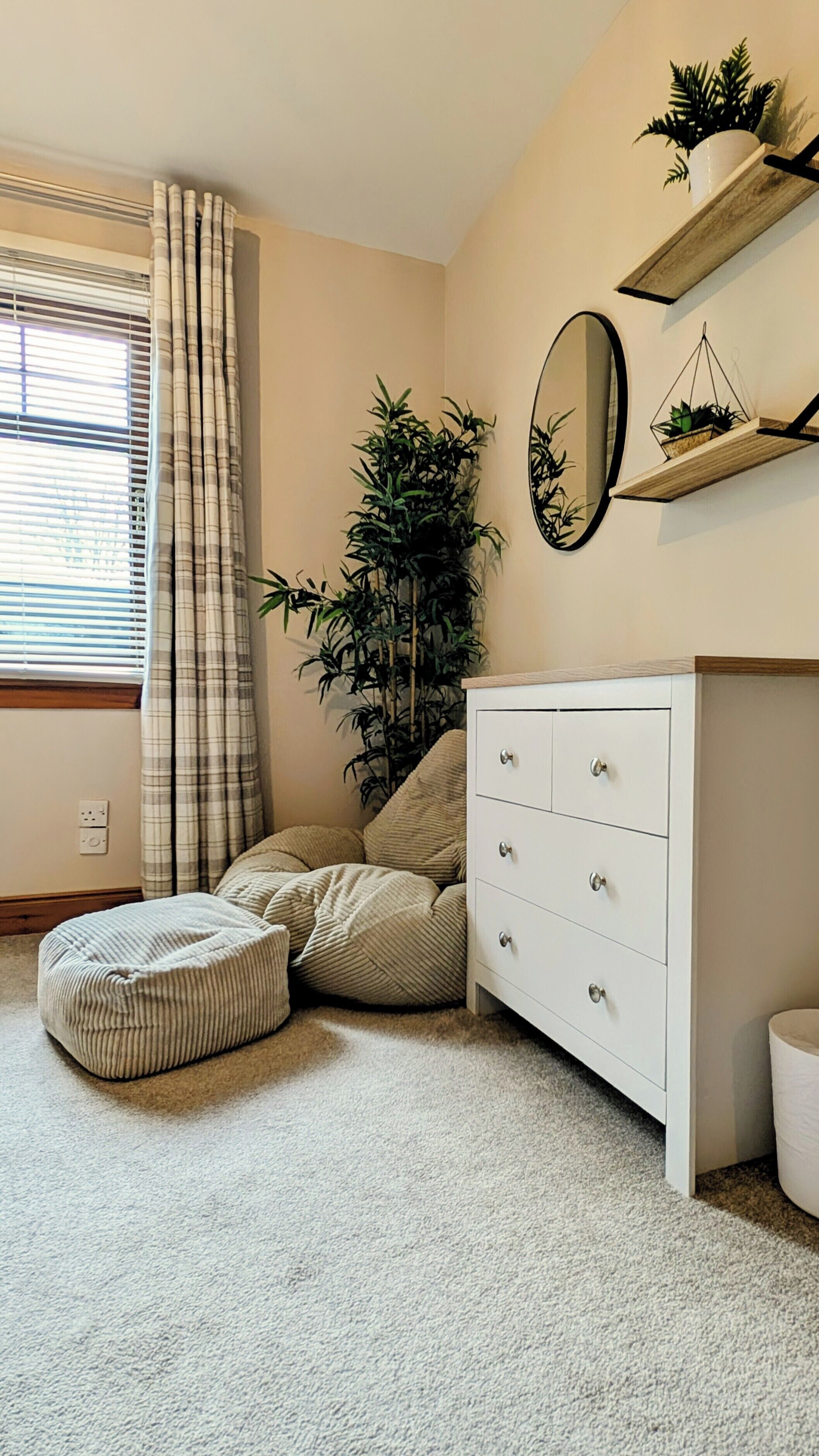 Een slaapkamer met een witte dressoir, een spiegel, een raam met zonwering, een potplant en een beigekleurig vloerkleed.